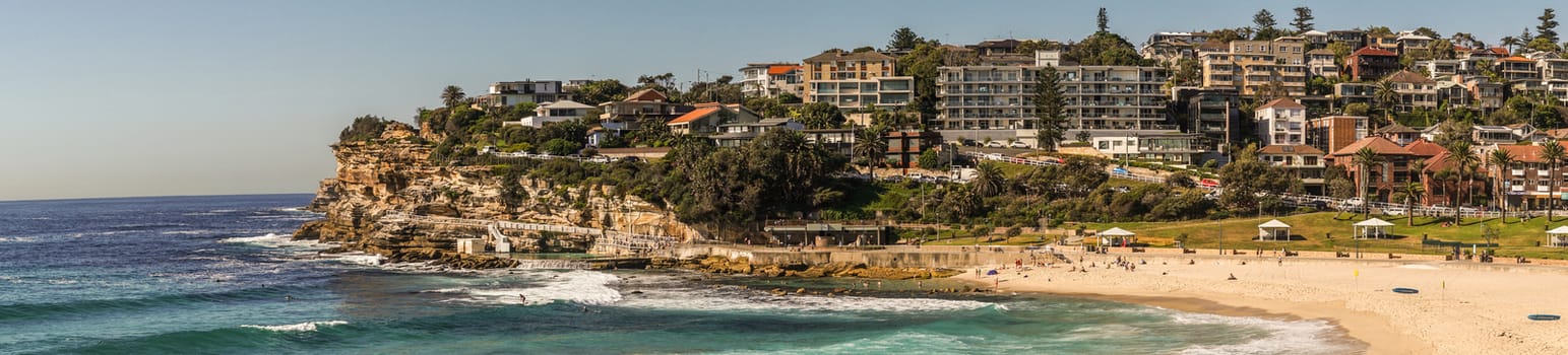 Sydney, Australia - February 11, 2019: Panorama shot of Bronte Beach, sea water, yellow sand, green park under blue sky, seen from North cliff. Band with housing on horizon. Brown rocks in foreground. People active.