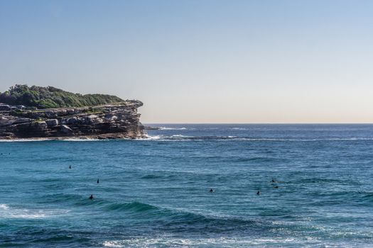 Sydney, Australia - February 11, 2019: Gray with green vegetation on top North Cliff point of Bronte Beach, blue sea water with swimmers, under light blue sky, seen from South cliff.