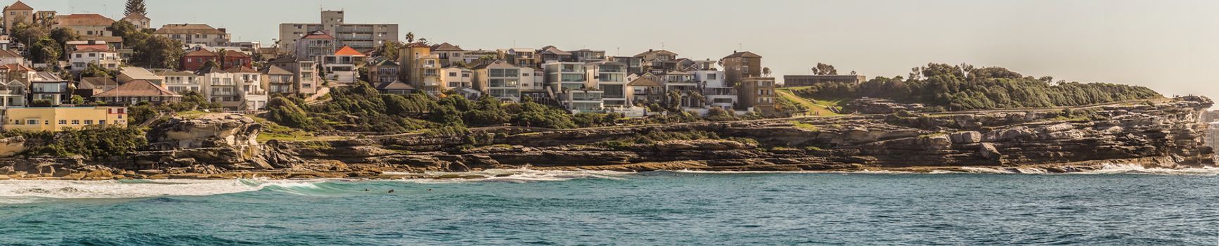 Sydney, Australia - February 11, 2019: Panorama shot of Bronte Beach, sea water under blue sky, seen from South cliff. Band with housing on horizon.