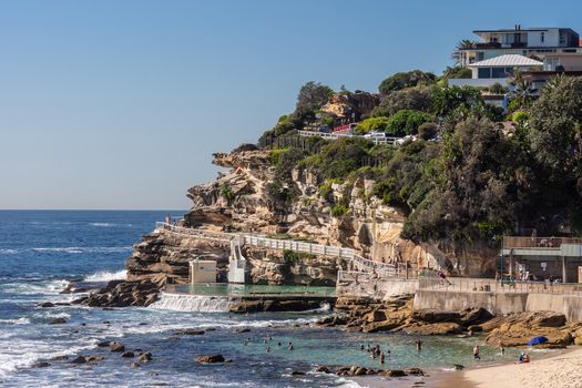 Sydney, Australia - February 11, 2019: Focus on the South Cliff at Bronte Beach with swimming pool in front. Peple in water and on sand. Housing in green vegetation on top of rocks.