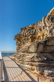 Sydney, Australia - February 11, 2019: Path ends at spectacular rock outcrop at Bronte Beach South cliffs, made by erosion.. Yellows and browns under blue sky. Sponge-like borders of shell like plates sticking out.