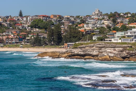 Sydney, Australia - February 11, 2019: Wide shot of Bronte beach with neighborhoods above and rocks to the north. Blue sea and blue sky. Waves crashing on rocks.