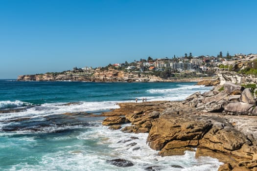 Sydney, Australia - February 11, 2019: Wide shot of Bronte beach with neighborhoods above and rocks to the north and south. Blue sea and blue sky. Waves crashing on rocks.