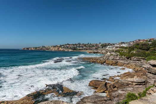 Sydney, Australia - February 11, 2019: Wide shot of Bronte beach with neighborhoods above and rocks to the north and south. Blue sea and blue sky. Waves crashing on rocks.