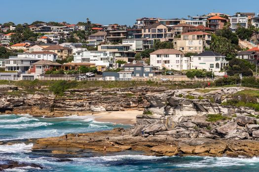 Sydney, Australia - February 11, 2019:  Tamarama beach with neighborhoods above and rocks to the north. Blue sea and blue sky. Waves crashing on rocks.