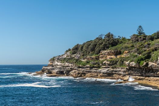 Sydney, Australia - February 11, 2019:  South side rocks and cliffs at Bondi beach. Green vegetation on top. People walking on path. Blue water and sky.