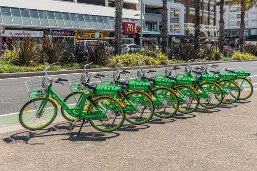 Sydney, Australia - February 11, 2019: Line of green and yellow Lime E-bicycles stationed on sidewalk of road bordering Bondi beach. People and cars.