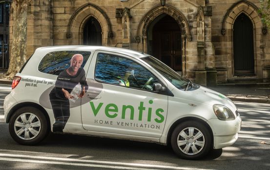 Sydney, Australia - February 12, 2019: Small white car with green advertisement and full picture of man for Ventis home ventilation systems. Back is yellow facade of Saint Patricks Church.