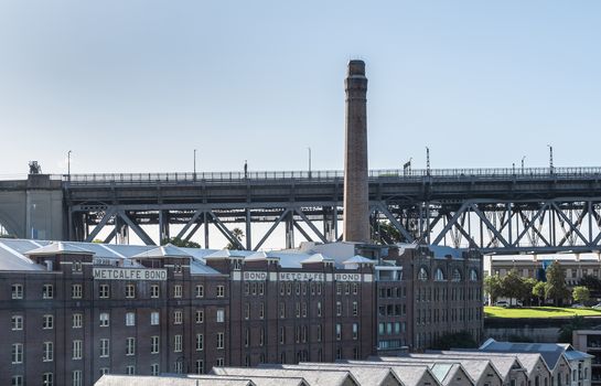 Sydney, Australia - February 12, 2019: Historic harbor warehouses with tall chimney sports Metcalfe Bond advertisement on facade. At foot of Harbour bridge.