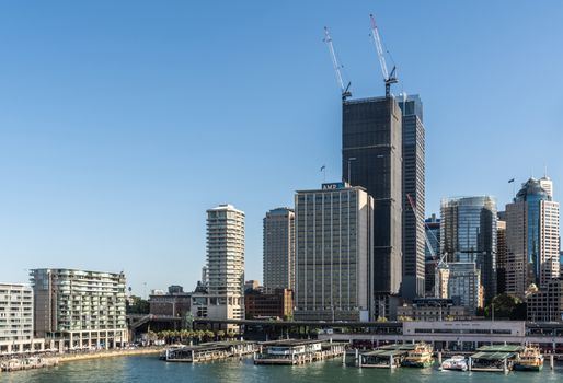 Sydney, Australia - February 12, 2019: Eastern side of ferry terminal and Circular Quay Railway Station plus skyline in back. Highrises under construction with cranes. Evening shot with light blue sky.