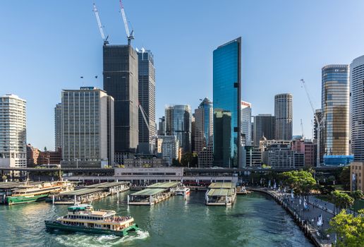 Sydney, Australia - February 12, 2019: Western side of ferry terminal and Circular Quay Railway Station plus skyline in back. Highrises under construction with cranes. Evening shot with light blue sky.