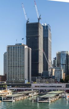 Sydney, Australia - February 12, 2019: Part of ferry terminal and Circular Quay Railway Station plus skyline in back. Highrises under construction with cranes. Evening shot with light blue sky.