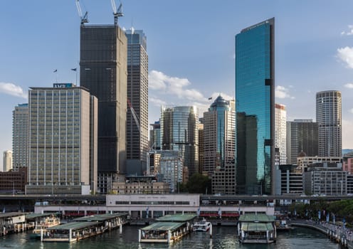 Sydney, Australia - February 12, 2019: Western Part of ferry terminal and Circular Quay Railway Station plus skyline in back. Highrises under construction with cranes. Evening shot with light blue sky.