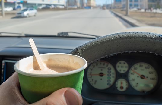 view of a glass of cocoa, chocolate, hot drink in the hand of a driver who is driving a car that is moving along a city street at speed