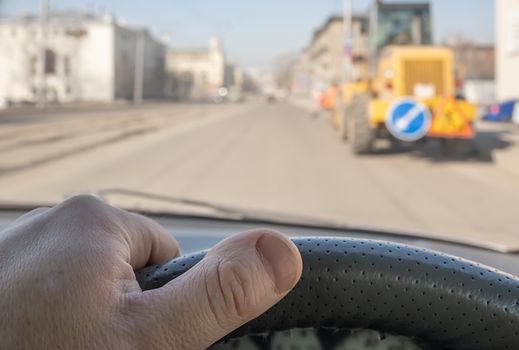 view of the driver hand on the steering wheel of a car passing through the courtyard of the street where the tractor stands, road signs and roadway repairs and maintenance are being carried out