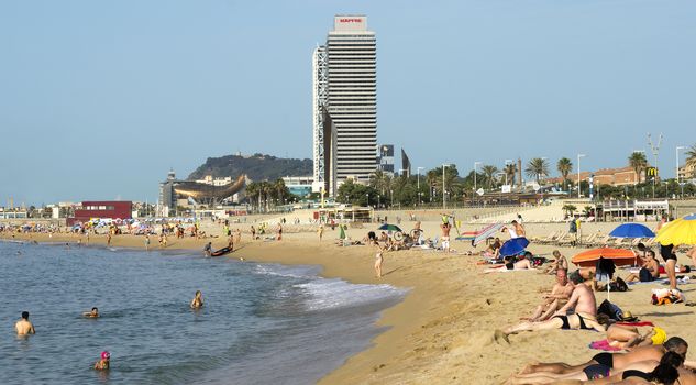 BARCELONA, SPAIN - JULY 5, 2015: Barceloneta Beach and skyscraper Torre Mapfre in the Olympic Port. It is named after its owner, Mapfre, an insurance company. This tower holds the title for highest helipad in Spain at 505 feet above ground.

Barcelona, Spain - July 5, 2015: Barceloneta Beach and skyscraper Torre Mapfre in the Olympic Port. It is named after its owner, Mapfre, an insurance company. This tower holds the title for highest helipad in Spain at 505 feet above ground. People are resting on the beach.