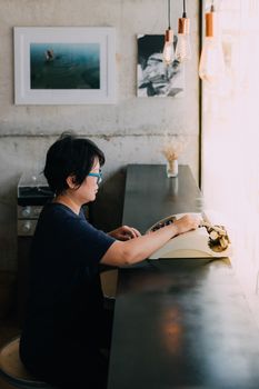 Asian woman 40s white skin with eyeglass sitting for writing something by vintage typewriter in a coffee shop cafe
