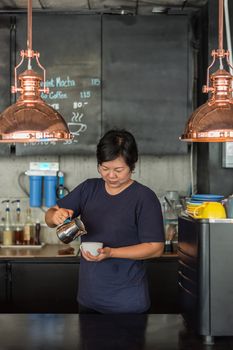 Asian woman 40s white skin work as a barista pouring latte froth to make a coffee latte art into the white coffee cup for serve to customers in the coffee shop cafe