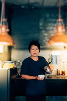 Asian woman 40s white skin work as a barista pouring latte froth to make a coffee latte art into the white coffee cup for serve to customers in the coffee shop cafe