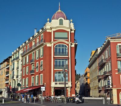 NICE, FRANCE - JUNE 5, 2014: The architecture of buildings on the Place Massena. The square was reconstructed in 1979. The square is bordered by red ochre buildings of Italian architecture.

Nice, France - June 5, 2014: The architecture of buildings on the Place Massena. The square was reconstructed in 1979. The square is bordered by red ochre buildings of Italian architecture. People are walking by street and are resting in the cafe.