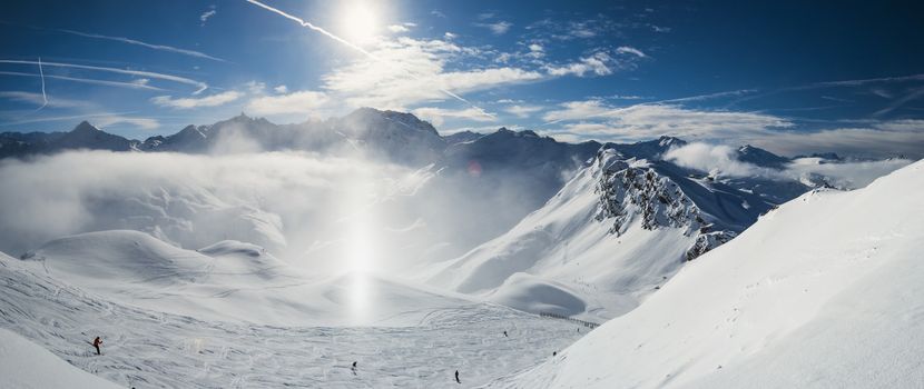 Panoramic view down snow covered valley in alpine mountain range with frost and sunlight sundog in air over ski piste