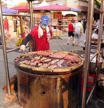 KAOHSIUNG, TAIWAN -- OCTOBER 15, 2016: An outdoor vendor cooks slabs of bacon, sausages, pork, eggs and rice in bamboo on a grill above a large barbecue pit.