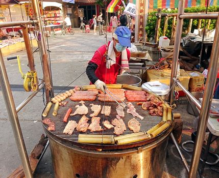 KAOHSIUNG, TAIWAN -- OCTOBER 15, 2016: An outdoor vendor cooks slabs of bacon, sausages, pork, eggs and rice in bamboo on a grill above a large barbecue pit.
