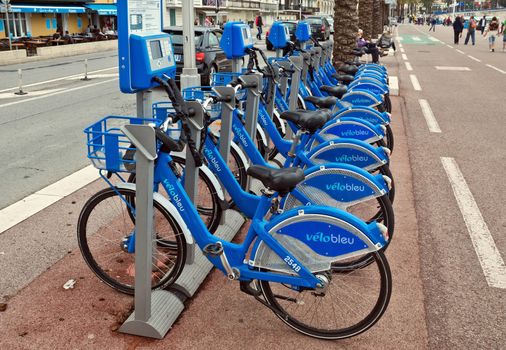 NICE, FRANCE - APRIL 27: Public Bicycles Sharing Station on April 27, 2013 in Nice, France. One of 120 stations in Nice. This service offers over 1200 self-service bicycles.

Nice, France - April 27, 2013: Row of public bikes for rent along the street. People are walking. Nice has 120 stations. This service offers over 1200 self-service bicycles. People are walking by Promenade.