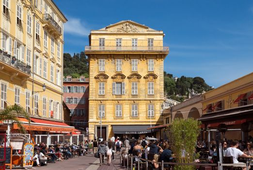 NICE, FRANCE - MAY 4: Old building and a cafe in the Cours Saleya on May 4, 2013 in Nice, France. The Cours Saleya is a place of outdoor restaurants and a market. 

Nice, France - May 4, 2013: Old building and a cafe in the Cours Saleya. The Cours Saleya is a place of outdoor restaurants, boutiques and a market. People are walking and relaxing.