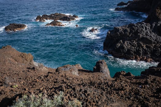 Volcanic rock formation, cliffs of black lava on the rocky shore with crushing white waves over the Atlantic Ocean. Blue sky background. La Palma, Canary Islands.