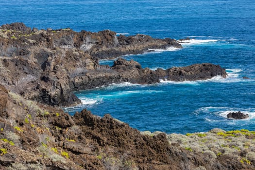 Volcanic rock formation, cliffs of black lava on the rocky shore with crushing white waves over the Atlantic Ocean. Blue sky background. La Palma, Canary Islands.