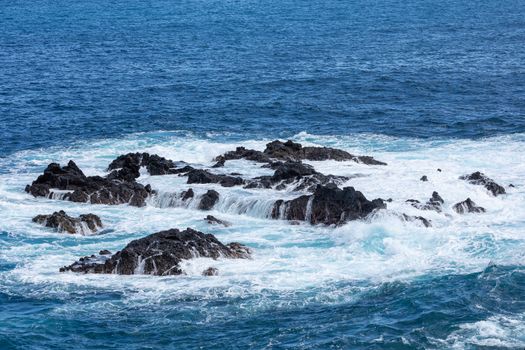 Volcanic rock formation, cliffs of black lava on the rocky shore with crushing white waves over the Atlantic Ocean. Blue sky background. La Palma, Canary Islands.