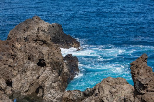 Volcanic rock formation, cliffs of black lava on the rocky shore with crushing white waves over the Atlantic Ocean. Blue sky background. La Palma, Canary Islands.