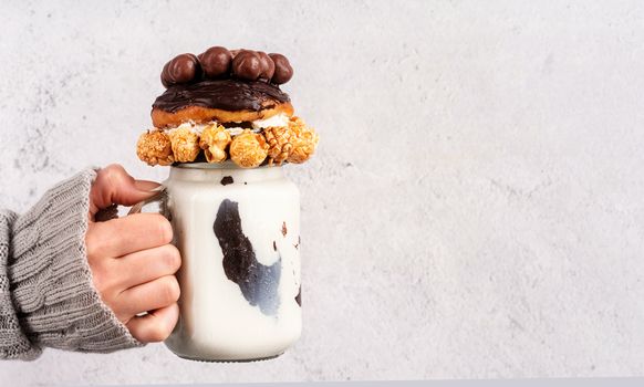 Female hand holding ice-cold chocolate donut milkshake in a mason jar decorated with caramel popcorn, whipped cream and cocoa candies front view with copy space