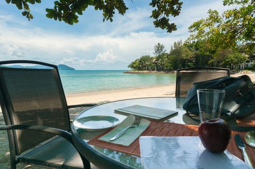 Shot of chairs and a table with healthy fruits on a beach with blue water and fluffy clouds. Shot in Malaysia shows a perfect vacation spot to relax in the summer