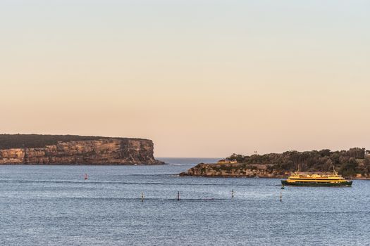 Sydney, Australia - February 12, 2019: North and South Head cliffs at gate between Tasman Sea and Sydney Bay during sunset. Cloudless pale sky. Gray water. Warm brown rocks and a yellow ferry boat.