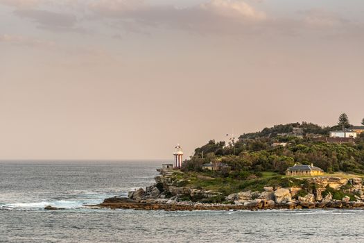 Sydney, Australia - February 12, 2019: South Head cliffs at gate between Tasman Sea and Sydney Bay during sunset. Cloudless pale sky. Gray water. Warm brown rocks and Hornby lghthouse.