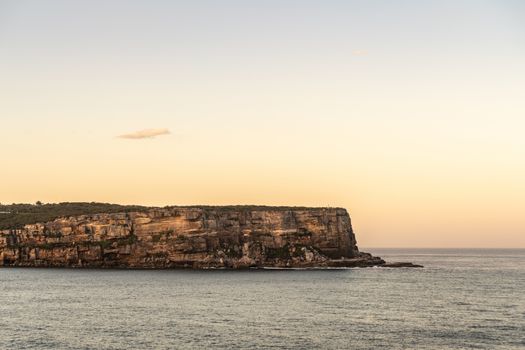 Sydney, Australia - February 12, 2019: North Head cliffs at gate between Tasman Sea and Sydney Bay during sunset. Almost cloudless pale sky. Gray water. Warm brown rocks.