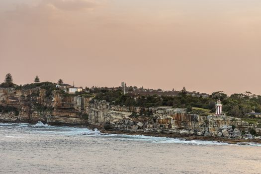 Sydney, Australia - February 12, 2019: South Head cliffs at gate between Tasman Sea and Sydney Bay during sunset. Cloudless orange sky. Gray water. Crashing waves. Warm brown rocks Hornby lighthouse.