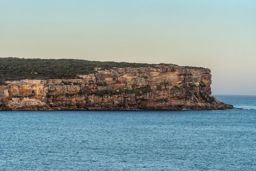 Sydney, Australia - February 12, 2019: North Head cliffs at gate between Tasman Sea and Sydney Bay during sunset. Almost cloudless pale sky. Gray water. Warm brown rocks.