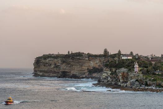 Sydney, Australia - February 12, 2019: South Head cliffs at gate between Tasman Sea and Sydney Bay during sunset. Cloudless pale sky. Gray water. Pilot boat. Crashing waves. Hornby lighthouse