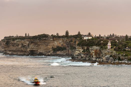 Sydney, Australia - February 12, 2019: South Head cliffs at gate between Tasman Sea and Sydney Bay during sunset. Cloudless pale sky. Gray water. Pilot boat. Crashing waves. Hornby lighthouse on cliff