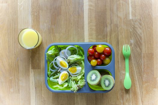 Collapsible silicon lunch box with food (salads, eggs, kiwi) on wooden table and orange juice