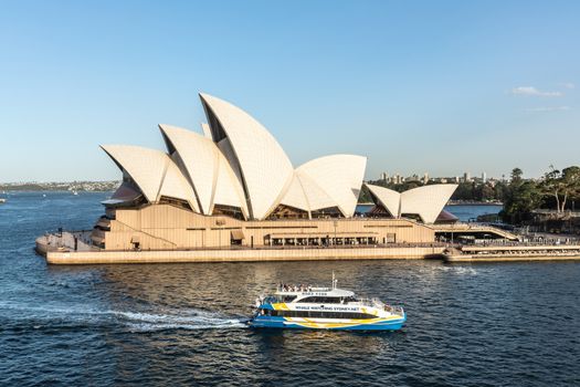 Sydney, Australia - February 12, 2019: Side view of the Opera House during sunset, with Whale Watching cruise vessel. Blue sky and water. Horizon is shore of bay.