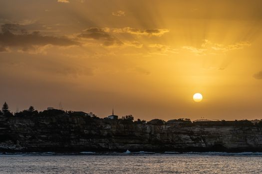 Sydney, Australia - February 12, 2019: Sunset over city skyline seen from Tasman Sea. Shoreline rocky cliffs. Yellow brown sky, sun rays and bow of Harbour bridge. 2 of 5.