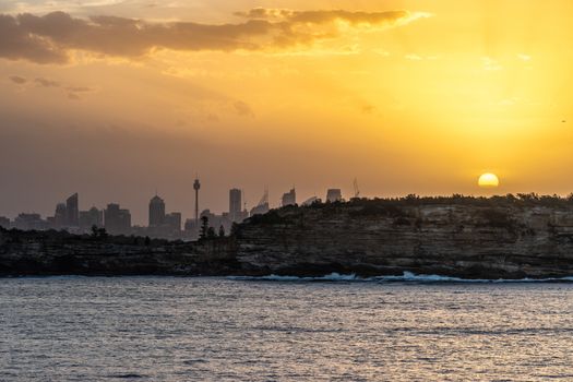 Sydney, Australia - February 12, 2019: Sunset over city skyline seen from Tasman Sea. Shoreline rocky cliffs. Yellow brown sky, sun rays. 3 of 5.