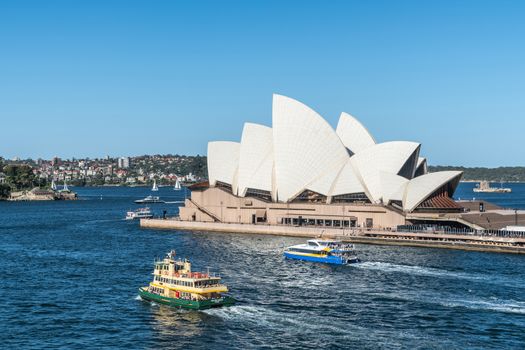 Sydney, Australia - February 12, 2019: Side view of the Opera House with ferries in front. Blue sky and water. Horizon is north shore of bay.