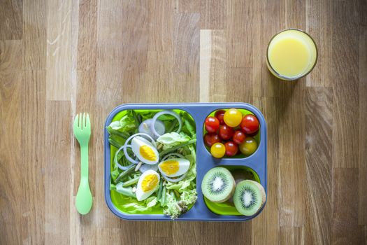 Collapsible silicon lunch box with food (salads, eggs, kiwi) on wooden table and orange juice