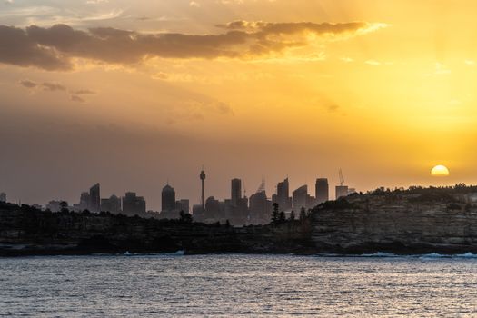 Sydney, Australia - February 12, 2019: Sunset over city skyline seen from Tasman Sea. Shoreline rocky cliffs. Yellow brown sky, sun rays. 4 of 5.