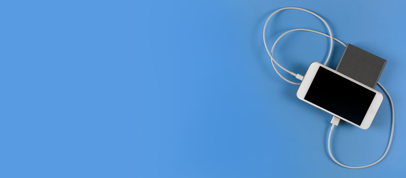 Top view smartphone charging with power bank on blue background with copy space. Smartphone charging with power bank by white USB cable.